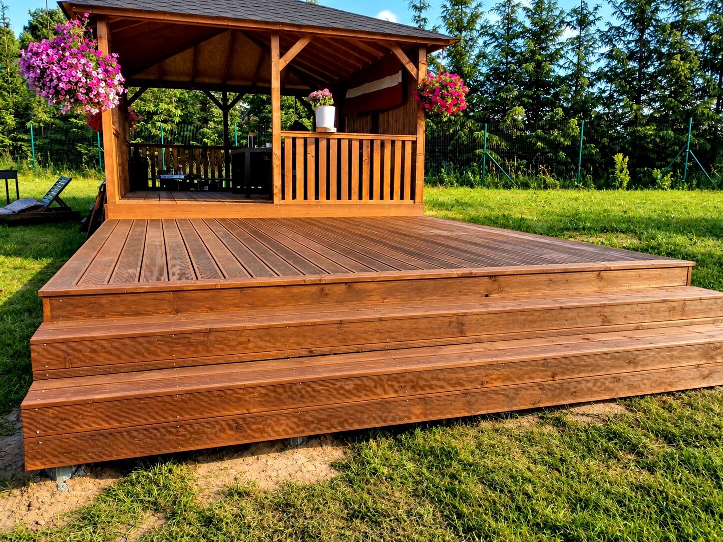A wooden terrace with steps leading up to a gazebo surrounded by greenery and flowers.