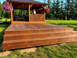 A wooden terrace with steps leading up to a gazebo surrounded by greenery and flowers.