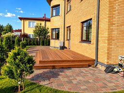 A wooden terrace is seen next to a brick house with a landscaped yard.